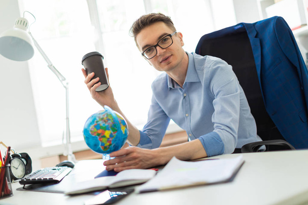 A young man sits in the office at a computer desk and holds a glass of coffee in his hand. A young man faces a globe. - Foto, afbeelding