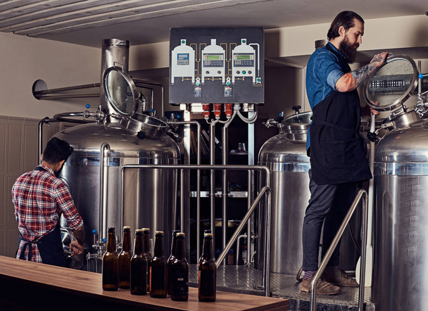 Два бородатых хипстера-мужчины в фартуке работают на пивоваренном заводе
. - Фото, изображение
