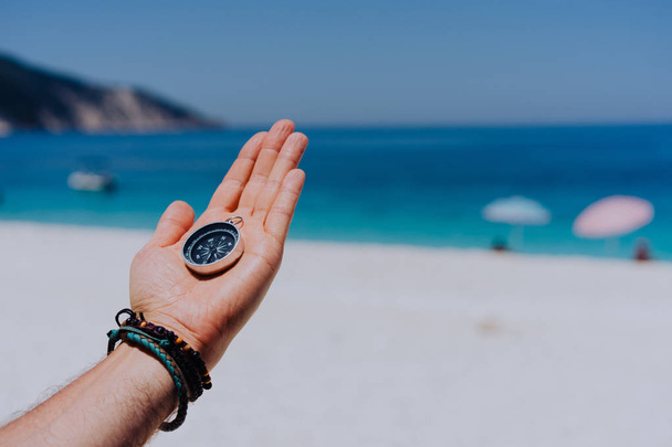 Stretched hand palm with black metal compass against sandy beach and blue sea. Follow your way, goal, wish concept. Point of view pov - Fotoğraf, Görsel