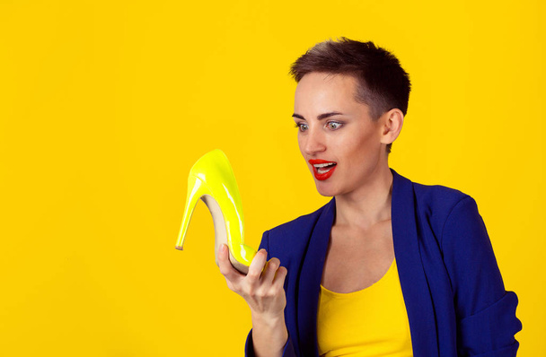 Woman holding looking at lime color shoe happy surprised. Women loves high heel shoes concept. Beautiful slim young happy female model isolated on yellow background. Model in shirt and blue suit. - Photo, Image