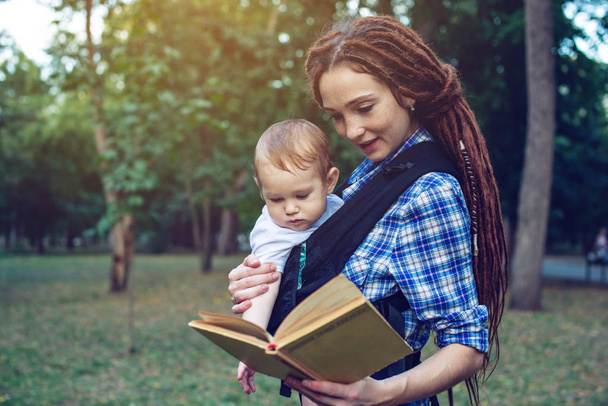 美しいママに赤ちゃんとエルゴ バックパックの公園でおとぎ話を読みます。家庭生活での子どもの教育 - 写真・画像