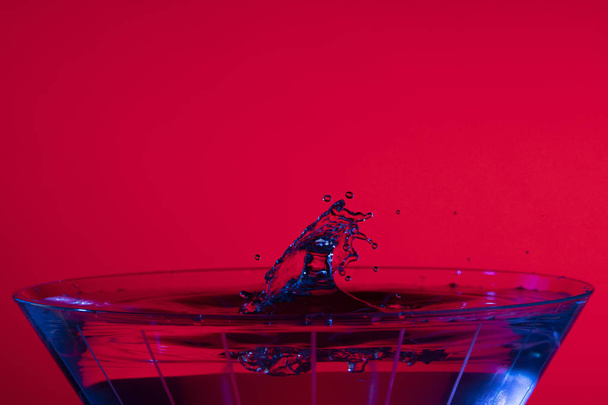 Droplets collide over a martini glass with red background and blue liquid highlights. - Foto, Imagen