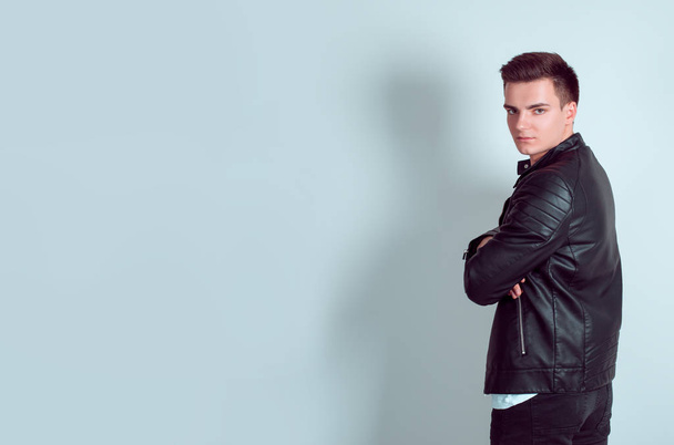 Portrait of confident young man with arms crossed turning back looking at you camera in studio isolated on light blue wall background Horizontal shot model in black leather jacket black jeans trousers - Photo, image