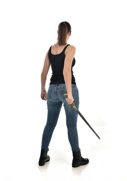 full length portrait of brunette girl wearing black single and jeans. standing pose, holding a sword. isolated on white studio background. - Photo, Image