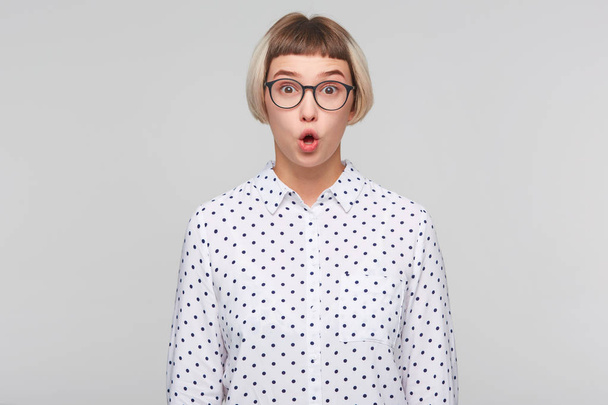 Closeup of surprised cute blonde young woman wears polka dot shirt and spectacles looks amusing and amazed isolated over white background - Photo, Image