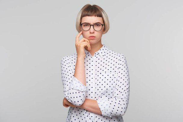 Close-up van peinzende geconcentreerde blonde jonge vrouw draagt polka dot shirt en bril houdt handen gevouwen en denken geïsoleerd op witte achtergrond - Foto, afbeelding