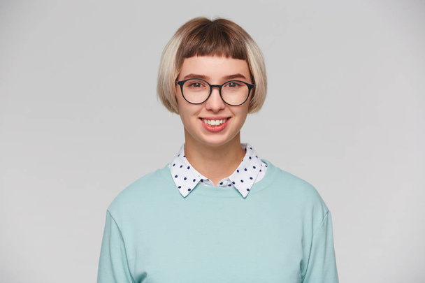 Primer plano de alegre atractiva joven mujer lleva sudadera azul y gafas se siente feliz, sonriendo y se ve directamente en la cámara aislada sobre fondo blanco
 - Foto, imagen