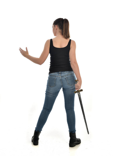 full length portrait of brunette girl wearing black single and jeans. standing pose, holding a sword. isolated on white studio background. - Photo, Image
