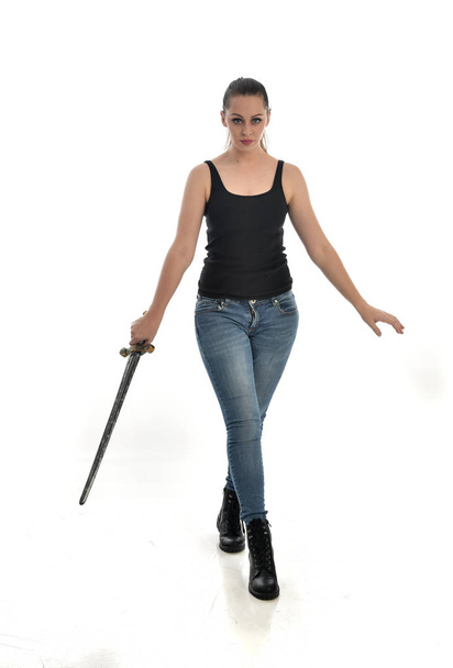 Ολόσωμο πορτρέτο μελαχρινή κοπέλα φορώντας το μαύρο ενιαίος και τζιν. όρθια στάση, κρατώντας ένα σπαθί. απομονωμένα σε φόντο λευκό στούντιο. - Φωτογραφία, εικόνα