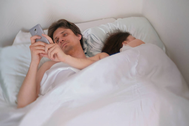 Husband is watching something in the mobile phone, while his wife is sleeping away. - Photo, Image