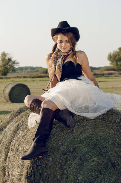 A young woman dressed as a cowgirl stands in a farmer's crop field corn at sunset and rests at a haystack - Photo, Image