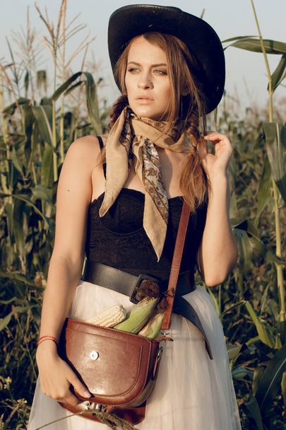A young woman dressed as a cowgirl stands in a farmer's crop field corn at sunset and rests at a haystack - Foto, Imagen