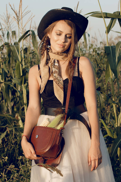 A young woman dressed as a cowgirl stands in a farmer's crop field corn at sunset and rests at a haystack - Foto, immagini