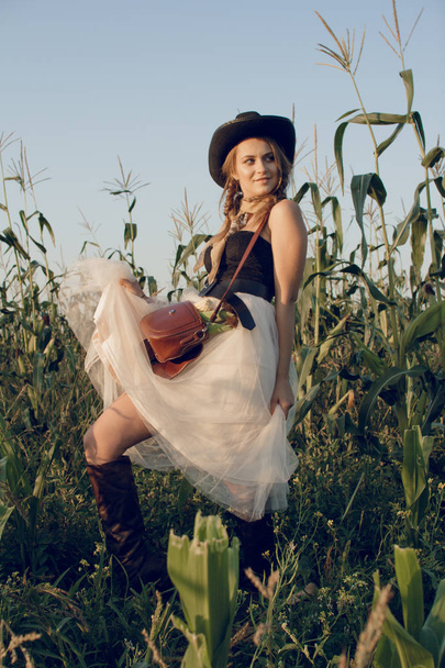 A young woman dressed as a cowgirl stands in a farmer's crop field corn at sunset and rests at a haystack - Foto, imagen