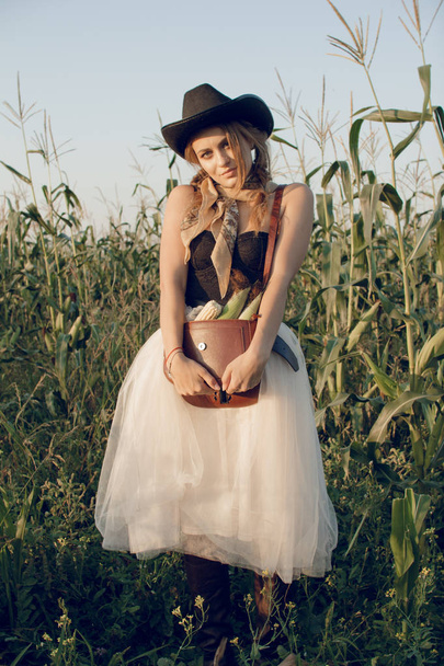 A young woman dressed as a cowgirl stands in a farmer's crop field corn at sunset and rests at a haystack - Foto, Imagen