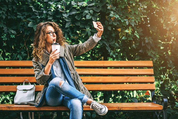 girl with curly hair taking selfie with coffee cup while sitting on bench - Photo, image