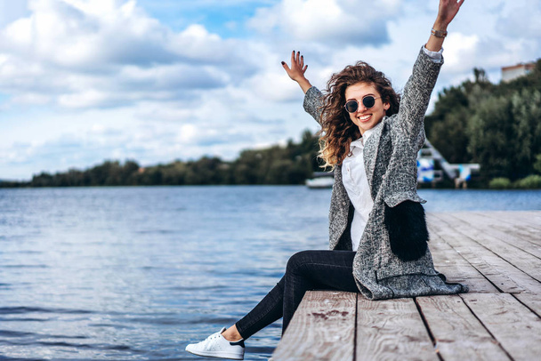 Pretty girl with curly hair posing with hands up near lake and smiling - Fotografie, Obrázek