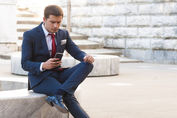 Joven hombre de negocios guapo en un traje y corbata consulta el teléfono inteligente sentado en un banco de mármol en el parque
 - Foto, imagen