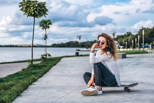 Pretty girl with curly hair sitting on skateboard in park and posing - 写真・画像