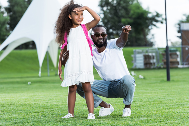 glimlachend Afrikaanse Amerikaanse vader naar dochter met school tas in park op iets - Foto, afbeelding