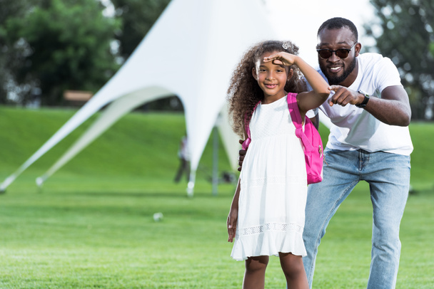 Αφρικανική αμερικανική πατέρα δείχνοντας στην κάμερα στην κόρη με σχολική τσάντα στο πάρκο - Φωτογραφία, εικόνα