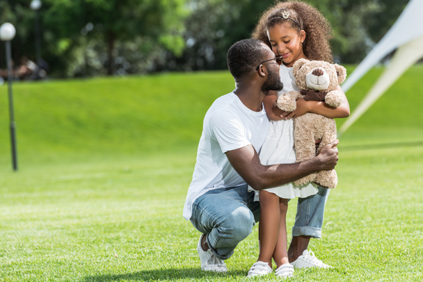 Αφρικανική αμερικανική πατέρα οκλαδόν και αγκαλιάζει αξιολάτρευτη κόρη με αρκουδάκι στο πάρκο - Φωτογραφία, εικόνα