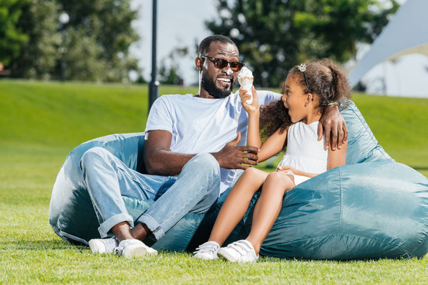 Αφρικανική αμερικανική κόρη διασκέδαση με παγωτό, ενώ κάθεται σε καρέκλες beanbag με τον πατέρα  - Φωτογραφία, εικόνα