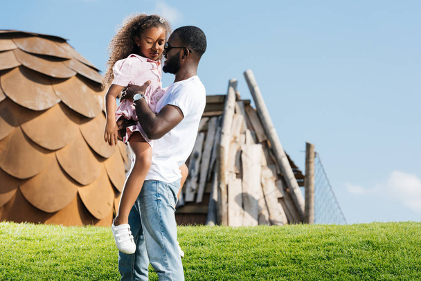Afrikaanse Amerikaanse vader bedrijf schattige dochter op groene heuvel op amusement park - Foto, afbeelding