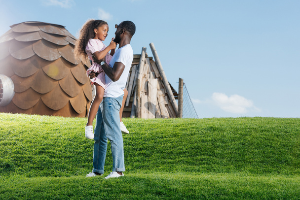 Африканский американский отец держит улыбающуюся дочь на зеленом холме в парке аттракционов
 - Фото, изображение