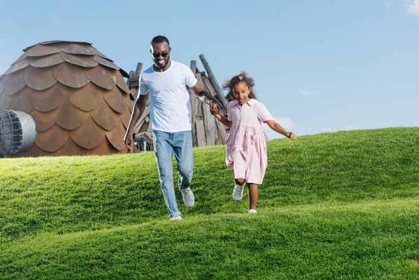 Αφρικανική αμερικανική πατέρας και κόρη, κρατώντας τα χέρια και να τρέχει στο λόφο στο λούνα παρκ - Φωτογραφία, εικόνα