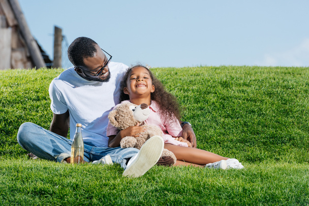 afro-amerikai apa és mosolygó lánya ül-dombon található a szóda és a mackó, vidámpark - Fotó, kép