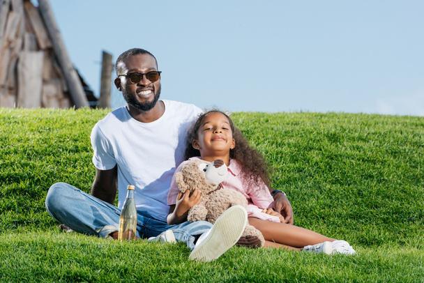 happy african american father and daughter sitting on hill with soda and teddy bear at amusement park - Photo, Image