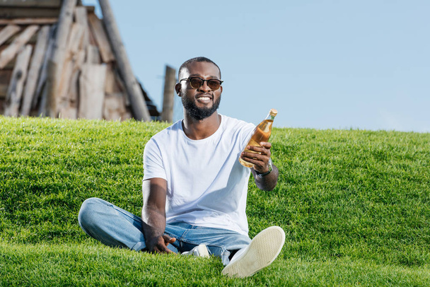 handsome african american man sitting on green hill and holding soda in glass bottle  - Photo, Image