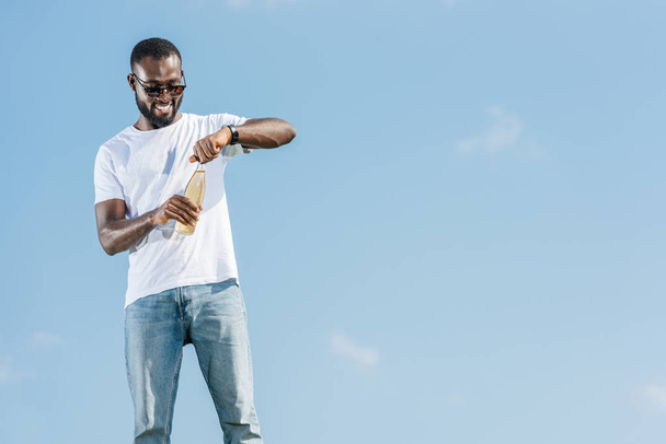 glücklich gutaussehender afrikanisch-amerikanischer Mann öffnet Limo-Flasche gegen blauen Himmel - Foto, Bild