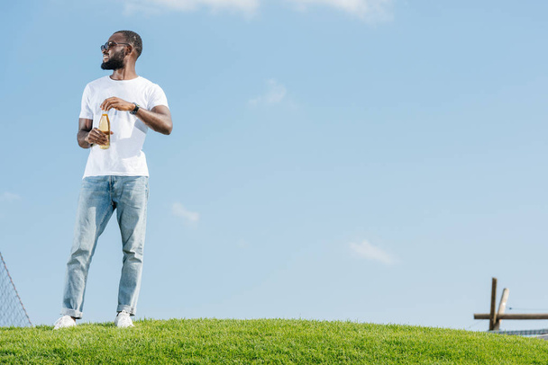 smiling handsome african american man opening soda bottle on green hill and looking away - Foto, Imagem