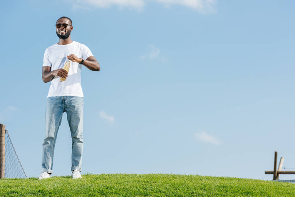 smiling handsome african american man opening soda bottle on green hill - Photo, Image