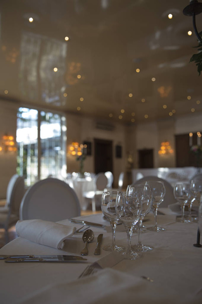 elegantly served with beautiful glasses, cutlery and decorated with flowers tables with white tablecloths and burning candles in candlesticks in the blurred background of the restaurant hall - Foto, Bild