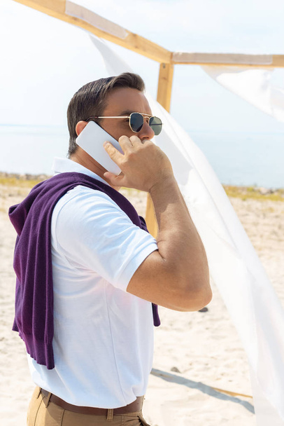 vista lateral del hombre con estilo en gafas de sol hablando en el teléfono inteligente en la playa de arena
 - Foto, Imagen