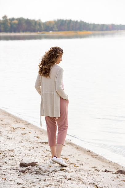 vista laterale di donna pensierosa con le mani in tasca in piedi sulla spiaggia di sabbia vicino al mare
 - Foto, immagini