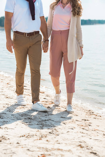 partial view of romantic couple holding hands while walking on sandy riverside - Foto, Imagen