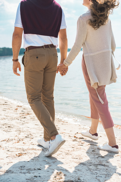 partial view of romantic couple holding hands while walking on sandy riverside - Fotó, kép