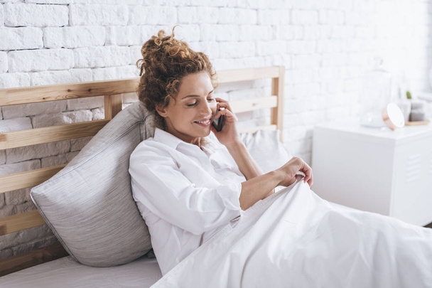 Довольно улыбающаяся белая женщина разговаривает по мобильному телефону, когда просыпается в своей постели
. - Фото, изображение