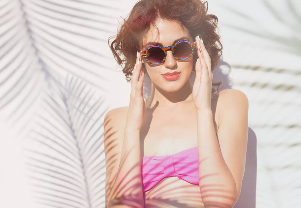 Woman in pink bra and sunglasses posing on background with shadows of palm leaves - Фото, изображение