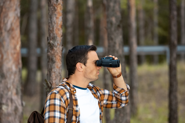 vista lateral do turista com mochila olhando através de binóculos na floresta
 - Foto, Imagem