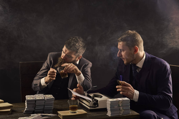 Men sitting at table with piles of money and typewriter. Illegal business concept. Businessmen discussing illegal deal while drinking and smoking, dark background. Company engaged in illegal business. - Foto, Imagem