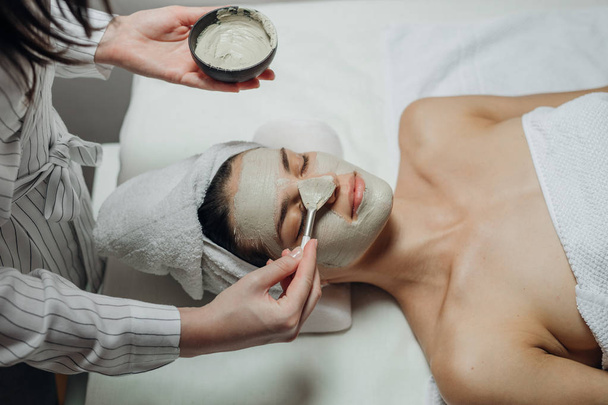 Caucasian cosmetologist woman applying a facial mask on a beautiful young Caucasian woman's face lying on the massaging bed. - Photo, image