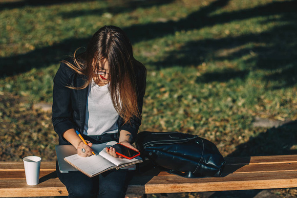 Young business woman working in the park, drinking coffee and browsing through her notebook. - Fotoğraf, Görsel