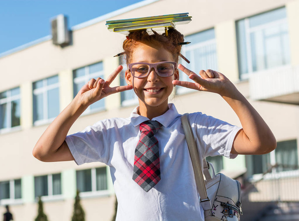 Ein Mädchen in Schuluniform, ein Buch auf dem Kopf und mit einer lustigen Frisur mit Buntstiften im Haar steht in der Nähe einer Schule - Foto, Bild