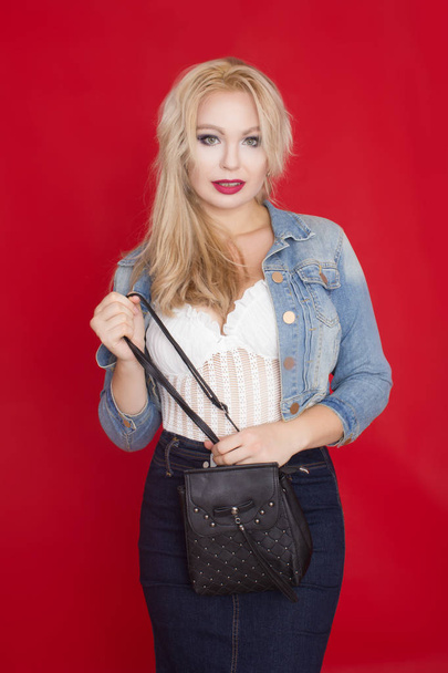 Sexy blonde woman in jeans skirt and jacket posing on red background with black bag in her hands. - Foto, immagini