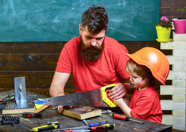 Father, parent with beard teaching little son to sawing with sharp handsaw, carpenter crafts. Fatherhood concept. Boy, child busy in protective helmet learning to use handsaw with dad - Photo, image